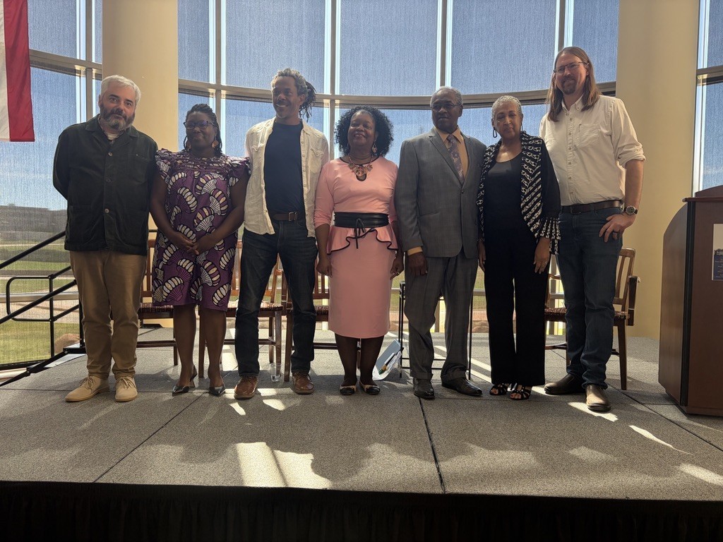 A group of people stands on a stage in front of a window, with a podium on the right.