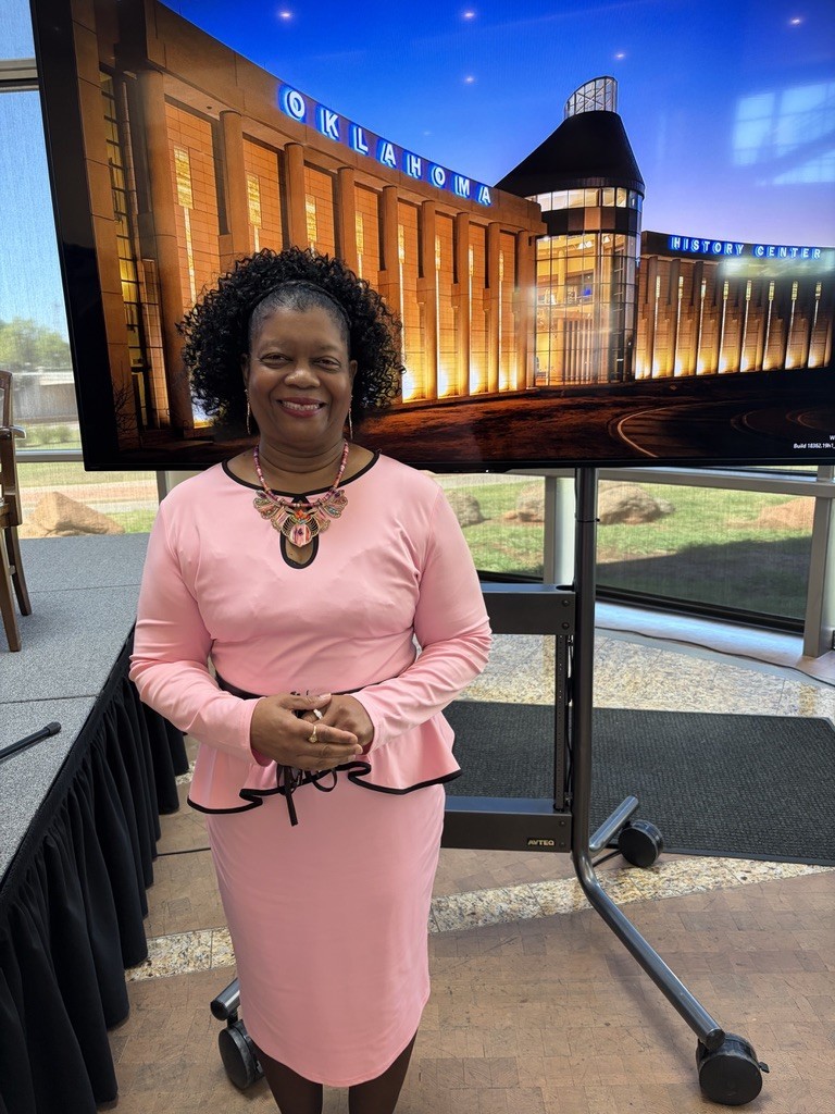 A woman in a pink dress stands before a large screen displaying a building, with a black backdrop and a tiled floor.