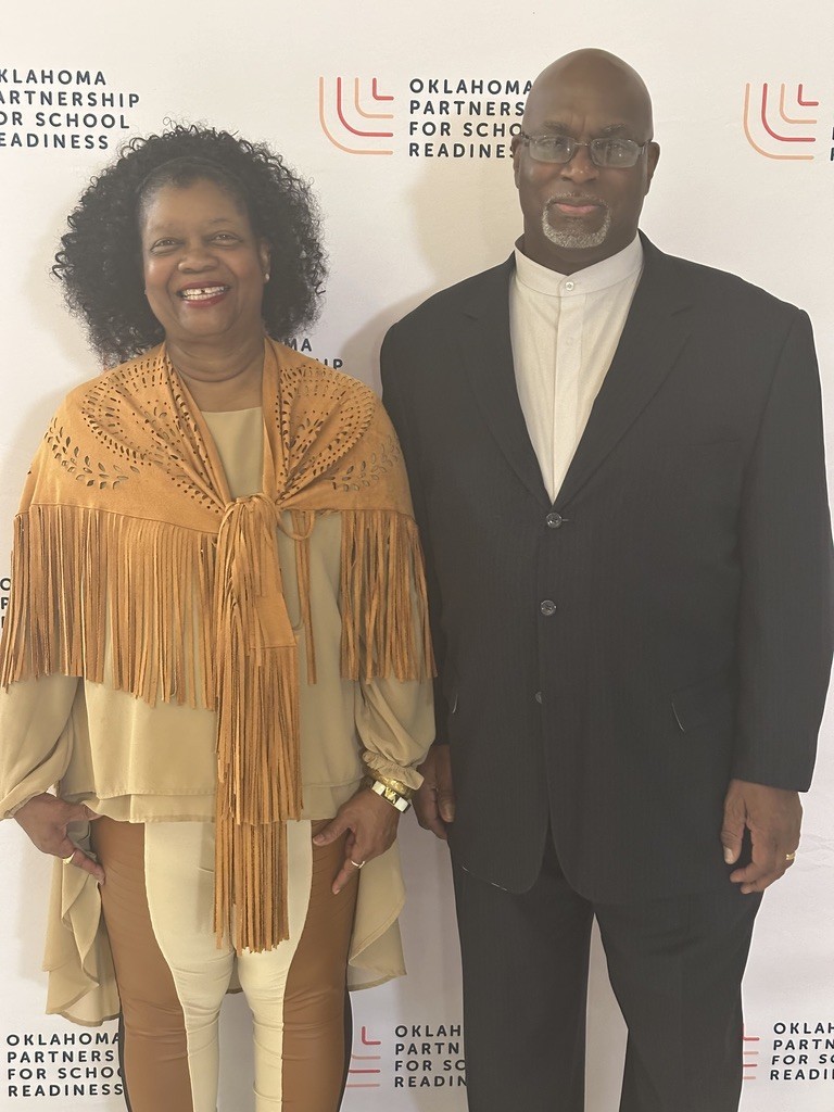Two individuals in formal wear pose for a photograph at an event with a banner that reads "Oklahoma Partnership for School Readiness".