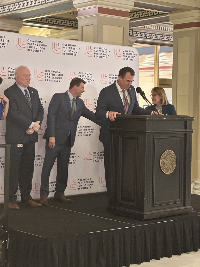 Four men and a woman stand on stage. A man speaks into a microphone. Four banners with logos are behind them.