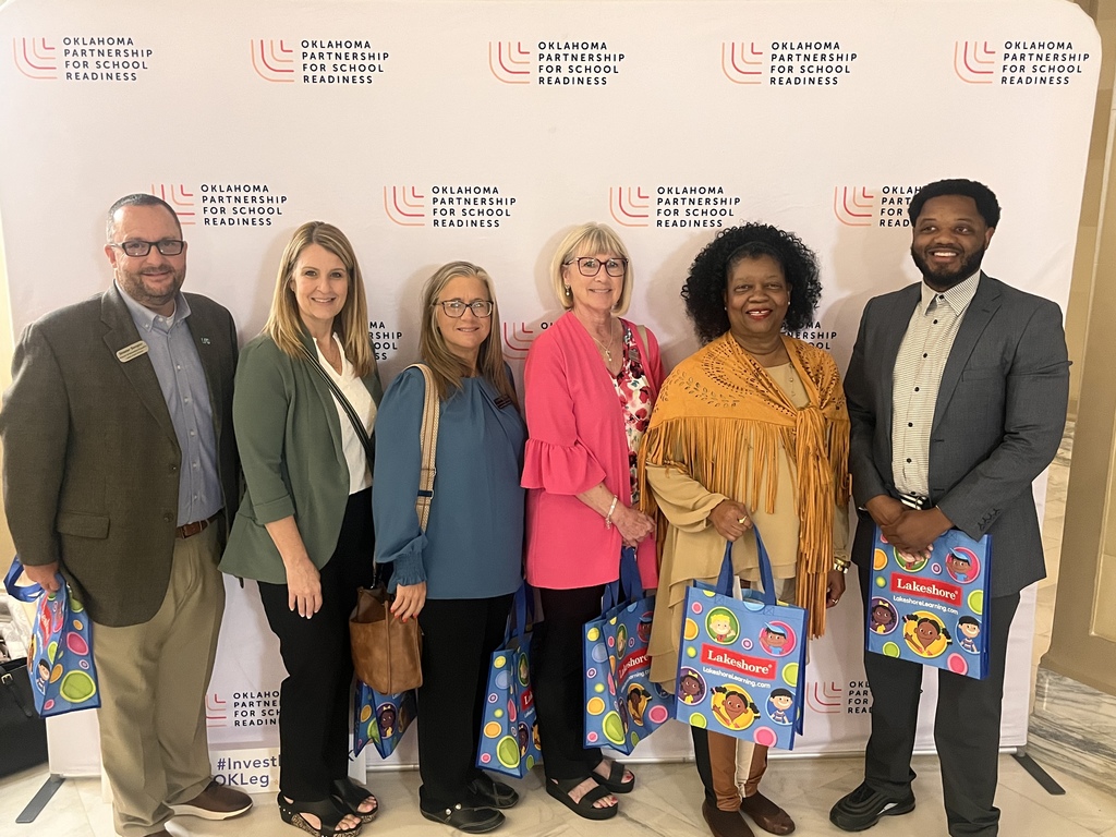 Five adults and one child pose in front of a wall with printed text, holding bags.
