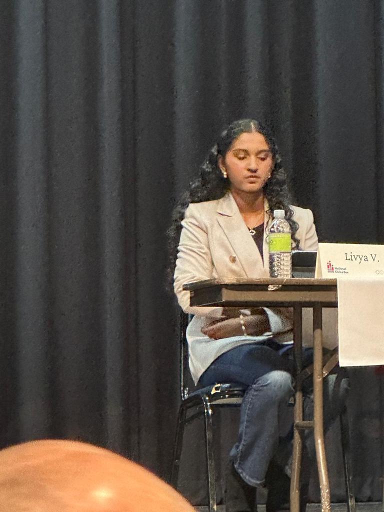 Woman seated at a table, looking down, wearing a white blazer, blue jeans, and black boots.