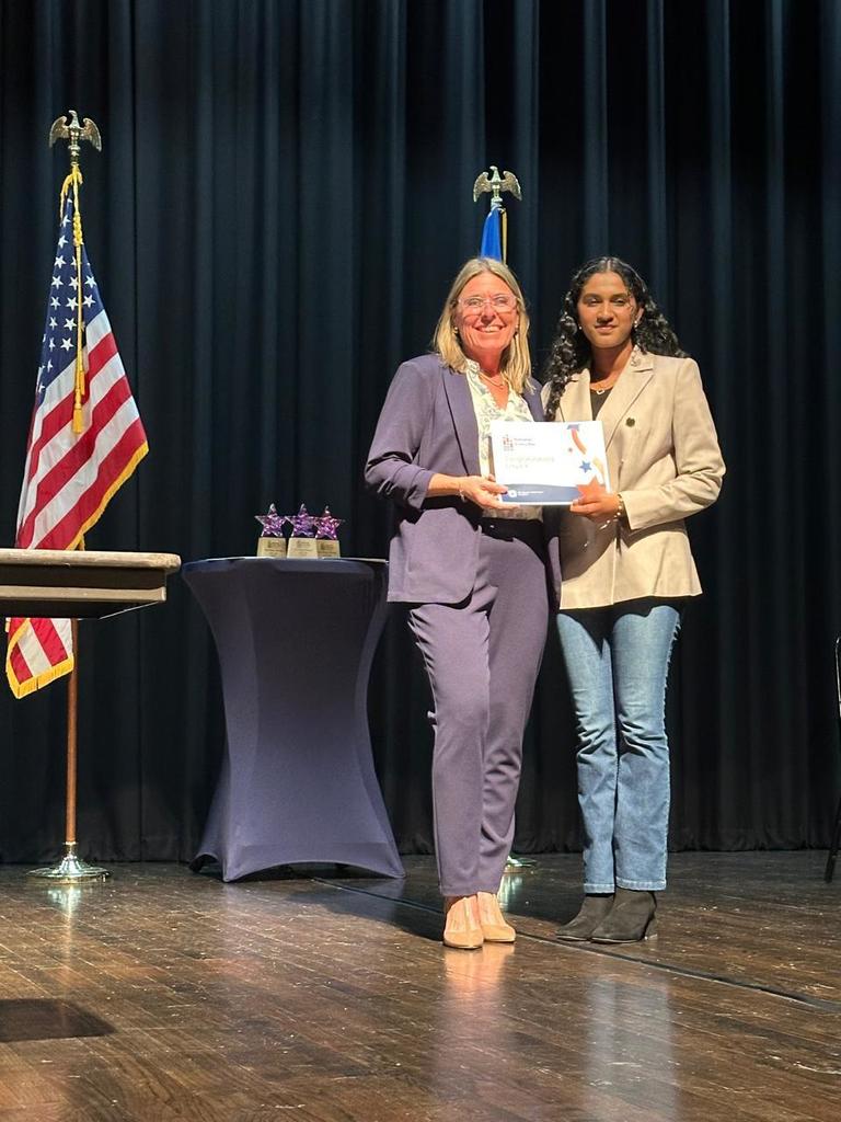 Two women stand on a stage, one holding a certificate. Behind them, a black curtain, a flag, and a small table.