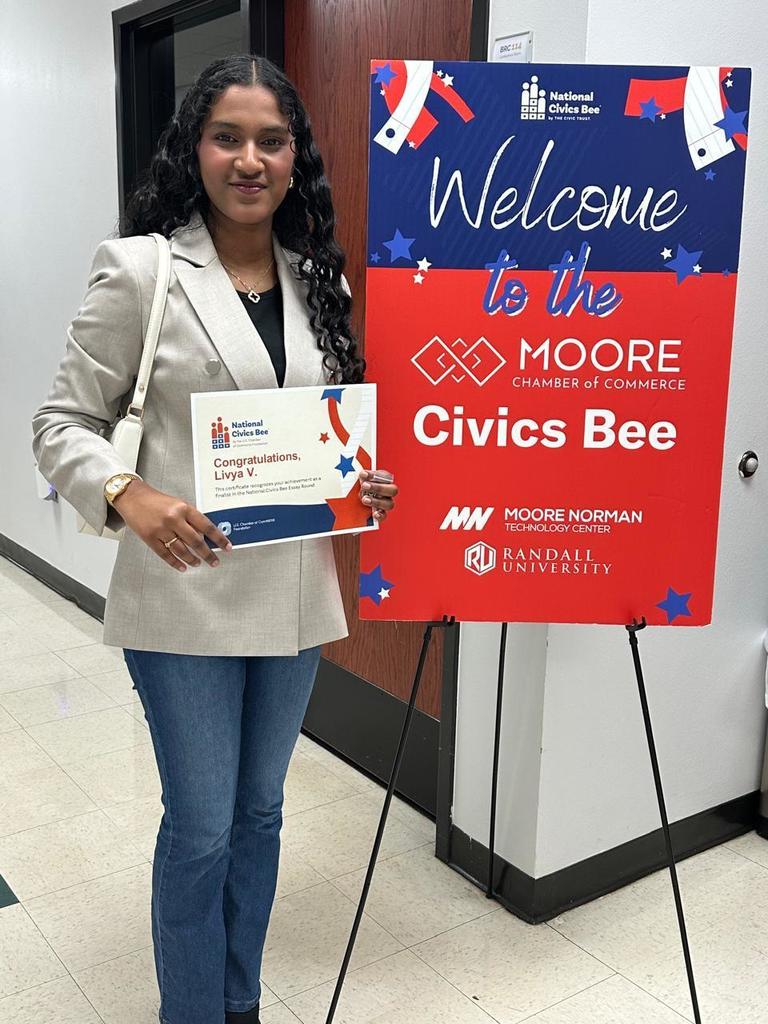 A woman in a light suit and jeans stands by a standee for Moore Civics Bee, holding a certificate.