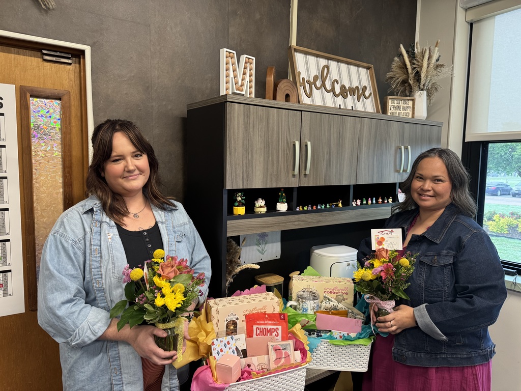 Two women stand, smiling, holding bouquets and gifts in a room with gray walls, cabinets, and a window.