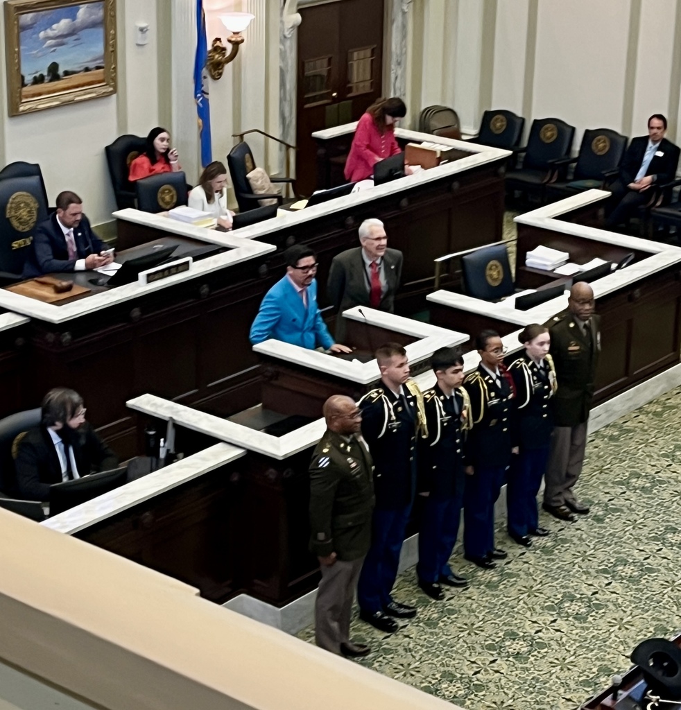 A legislative meeting with seated officials, a woman in red, and six uniformed individuals standing in front of the chairs.
