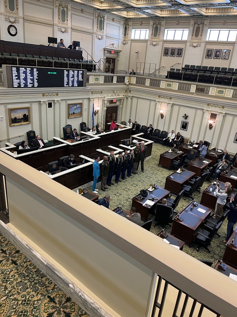 An indoor chamber with a high ceiling and multiple rows of tables and chairs. Several people are standing in a row.