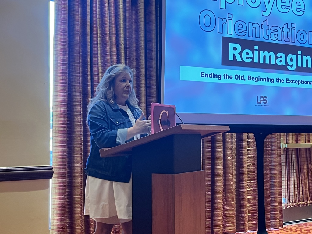 A woman speaks at a podium. A screen behind reads "Employee Orientation Reimagining" with text below.