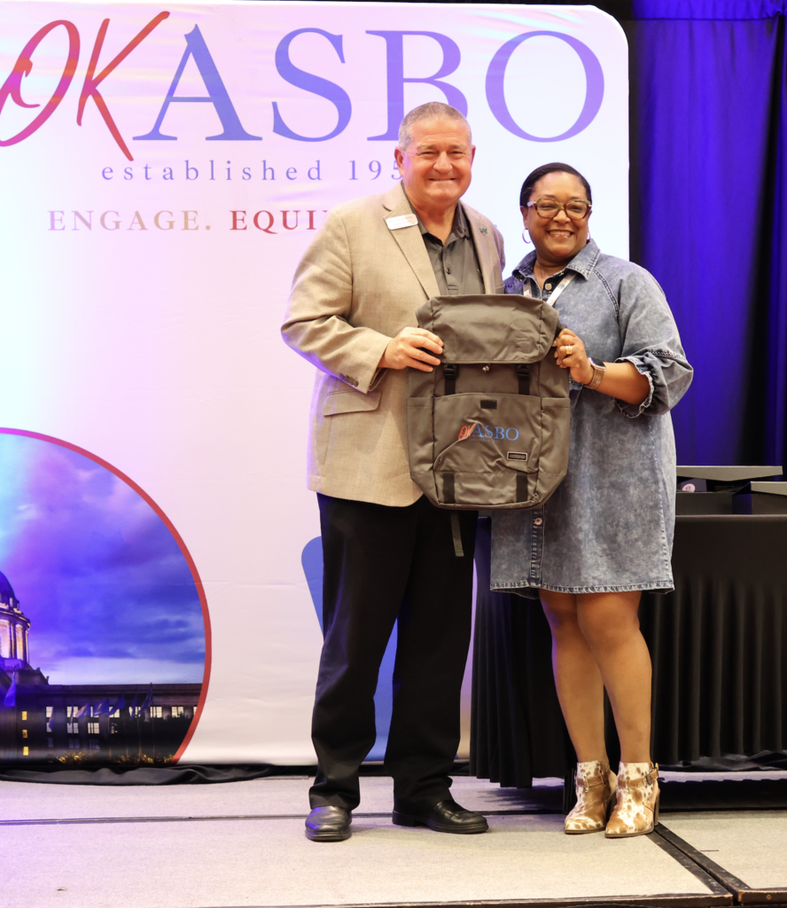 A man and woman stand on a stage holding a backpack. Behind them, a banner reads "OKASBO established 1973 ENGAGE. EQUIP."