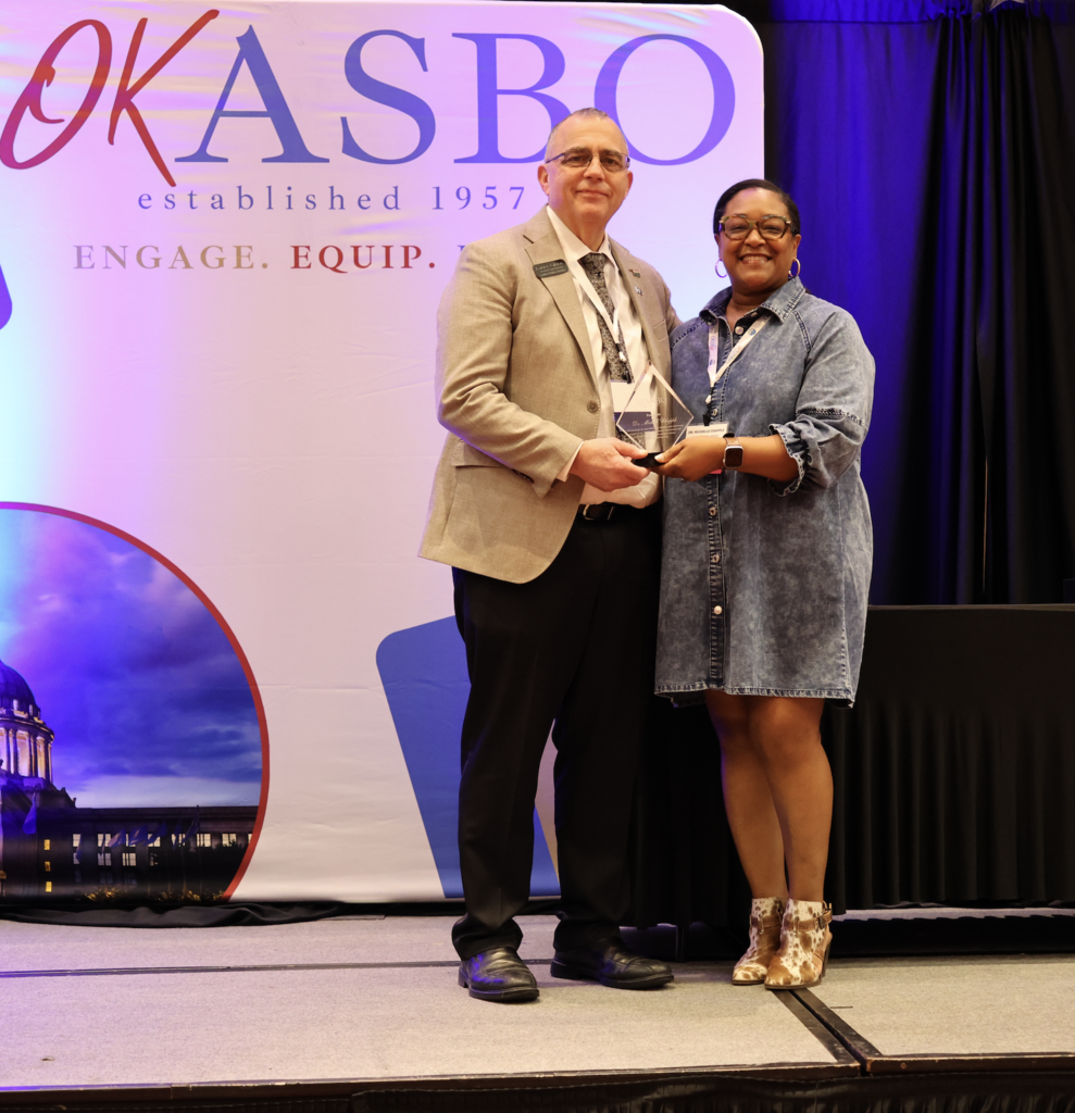 A man and woman stand on stage, holding awards. Behind them is a backdrop with text "OKASBO established 1957".