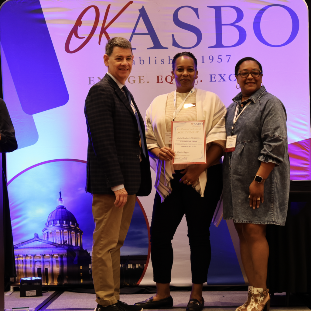 Three people stand on a stage, one holding a certificate. The man wears a suit and tie. The others wear a white jacket and a denim dress.