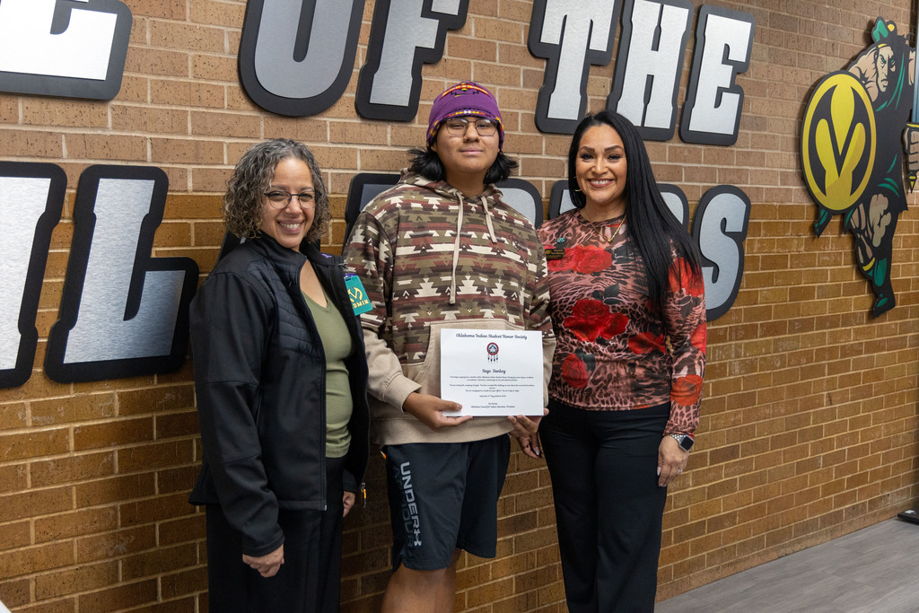 Three individuals pose for a photo in front of a brick wall. One holds a certificate.