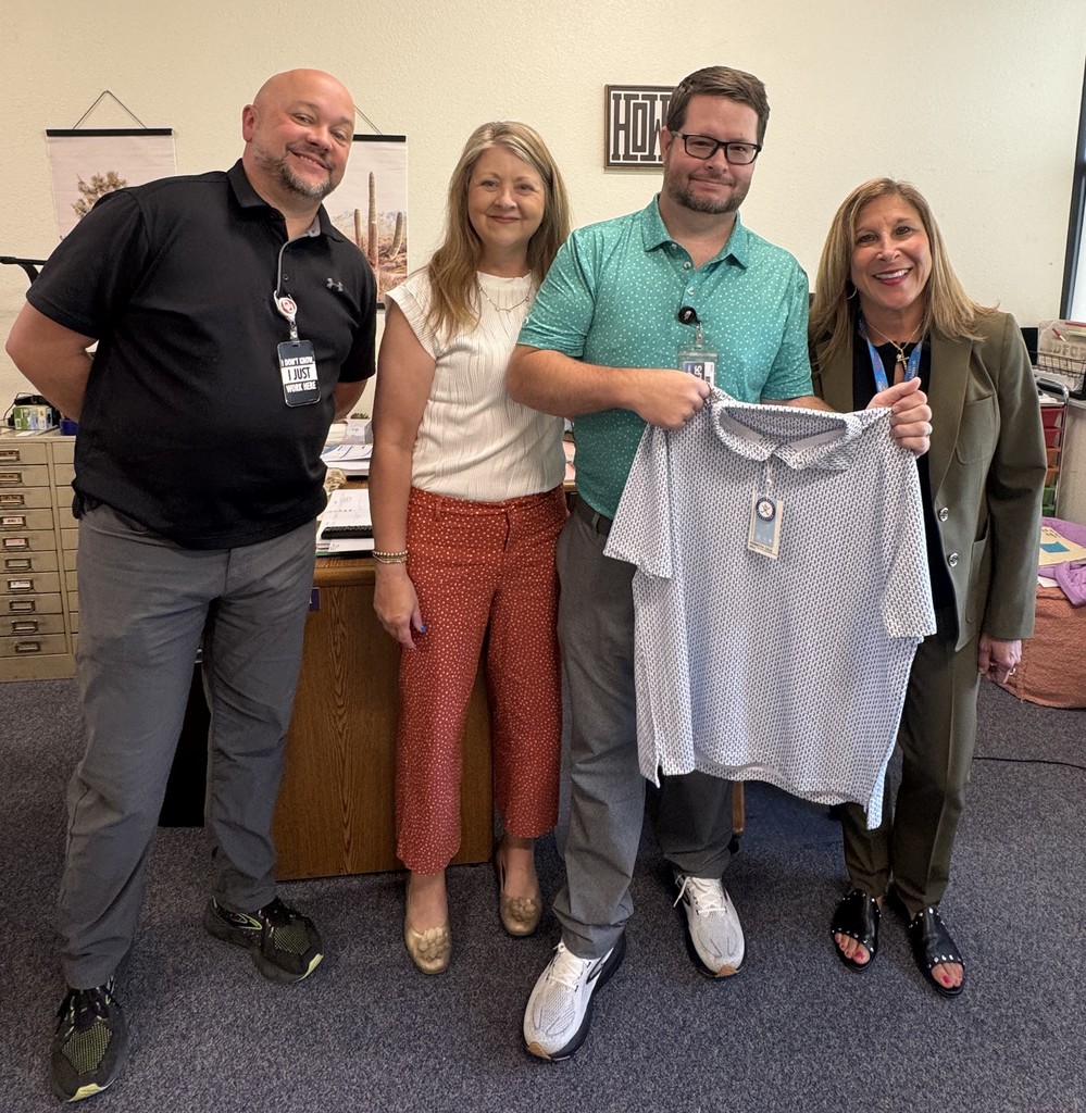 Four people standing in an office holding a white shirt and smiling at the camera.
