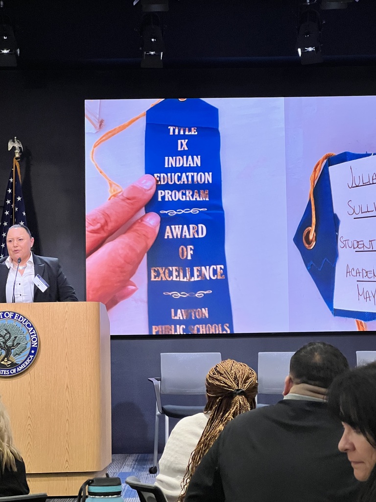 A podium with a person speaking at a ceremony. A large screen displays "Award of Excellence" and "Indian Education Program". Audience seated, partially visible.