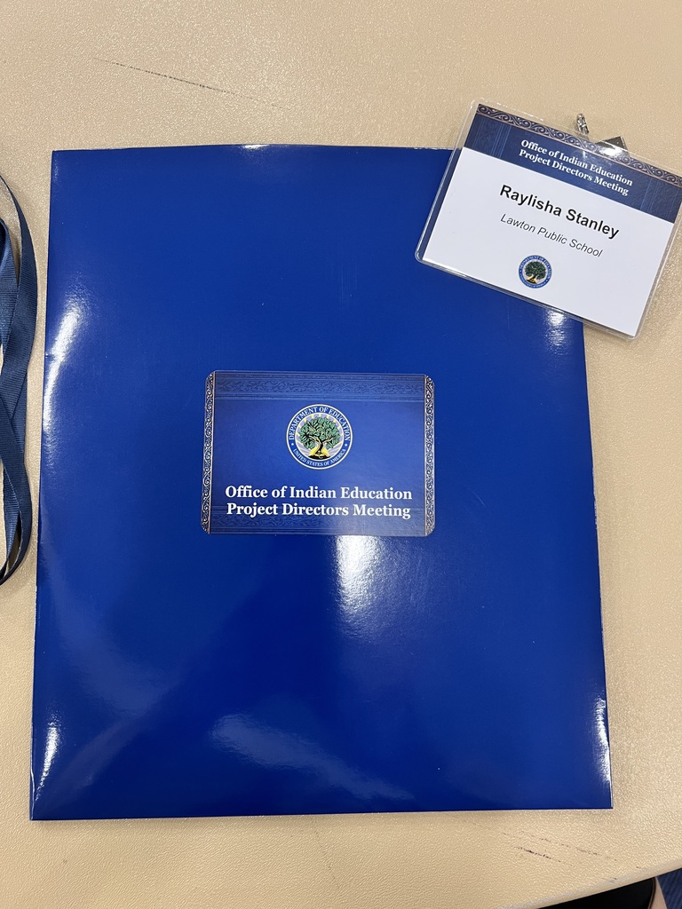 Blue folder with silver logo and "Office of Indian Education Project Directors Meeting" text. A white badge with "Baylsha Stanley" and "Leader" text is on the folder.