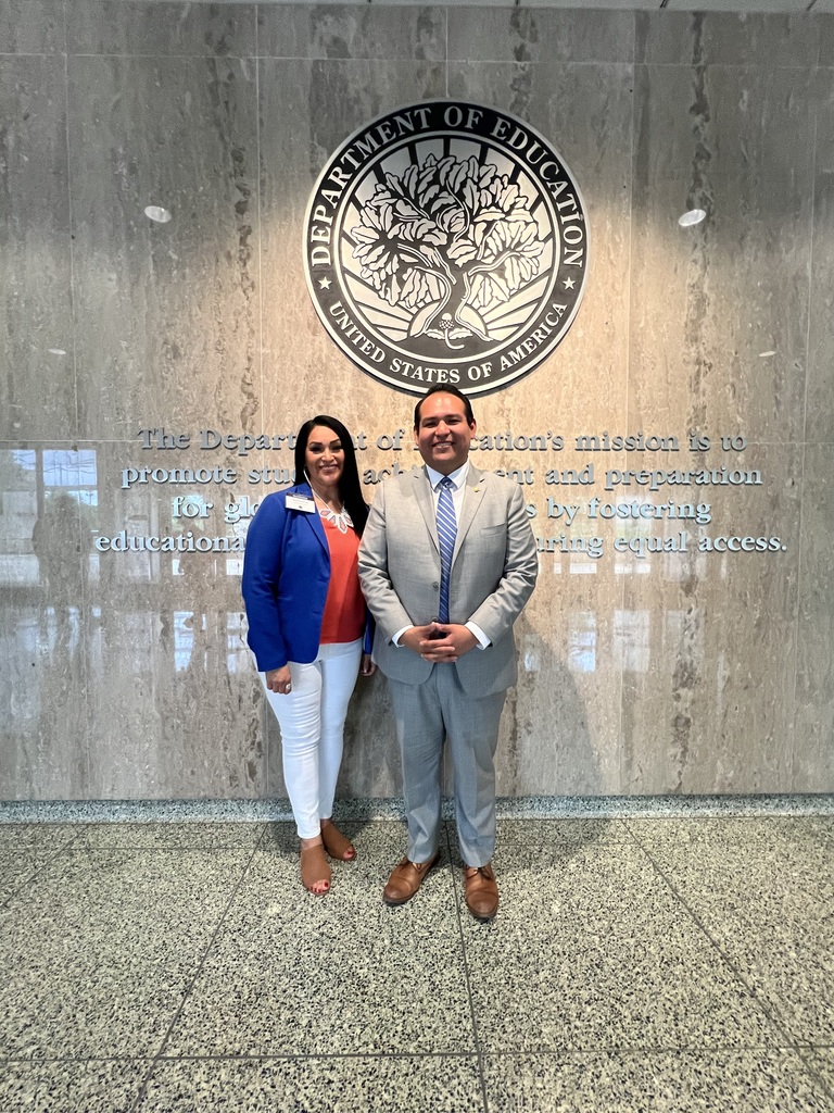 Two people stand in front of a concrete wall with a large logo of the Department of Education.