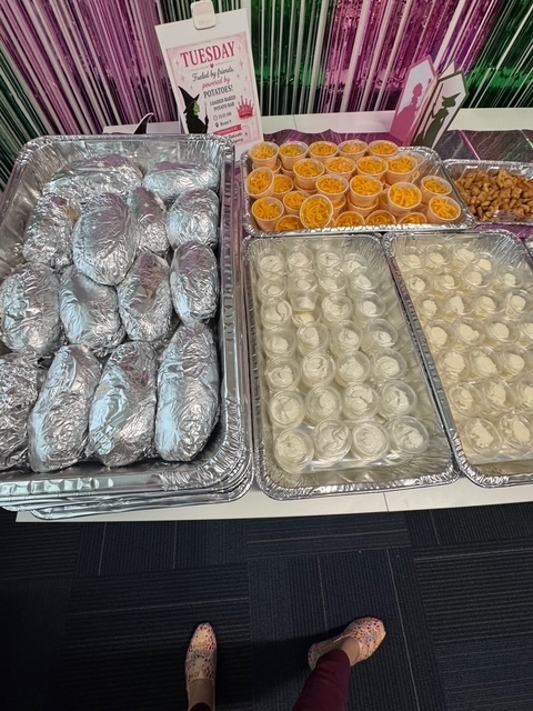 Trays of prepared food on a table with pink curtains in the background. Items include meat, cheese, and fruit.