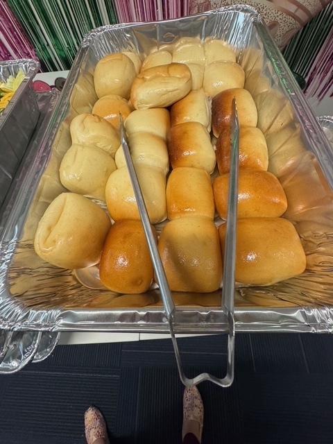 Tray with baked rolls, some golden brown. Metal tongs on the left. Background includes colorful drapes.