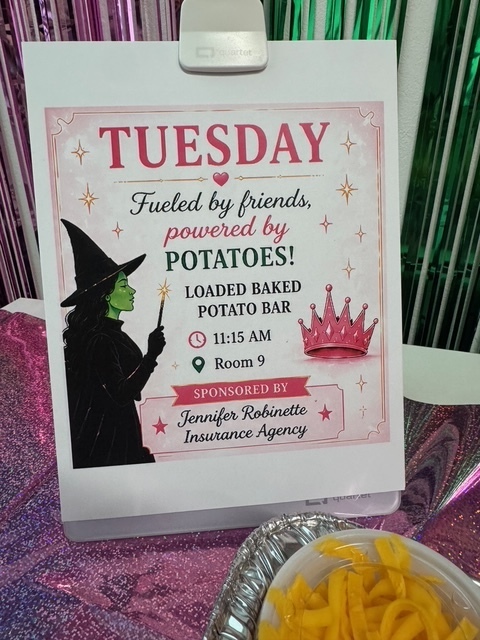A table holds a silver tray with a bowl of yellow food. A sign with "Tuesday" and a witch reads "Fueled by friends, powered by potatoes!" It also includes details about a potato bar.