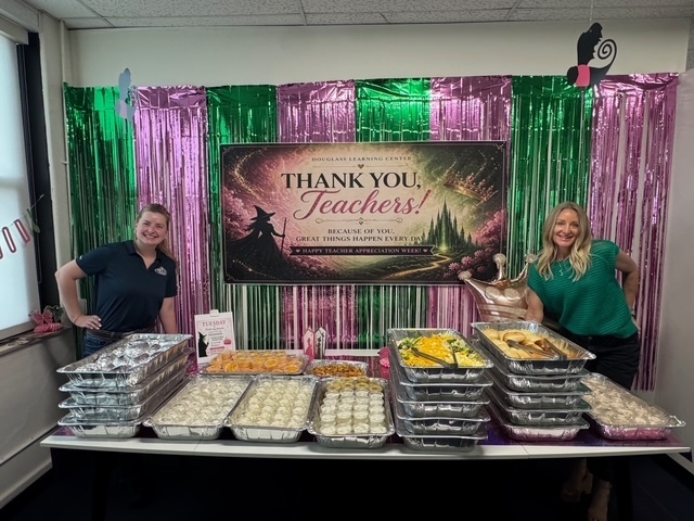 Two people behind a table, smiling. Banner behind says "Thank You, Teachers!" Food on table.