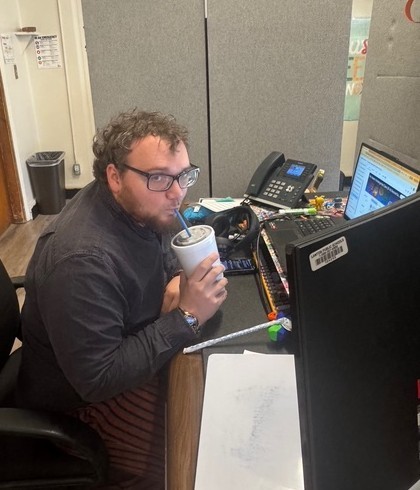 A person sits at a desk with a computer and drinks from a cup while wearing glasses.
