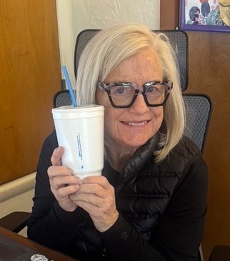 A woman with glasses sits in a chair, smiling, and holding a cup with a blue straw.