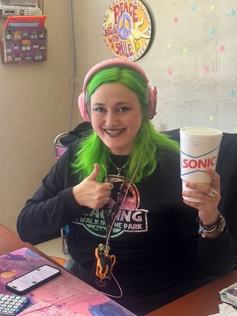 Woman with green hair wearing headphones, holding a Sonic Drive-In cup, smiling. A phone is on a table with a colorful tablecloth.