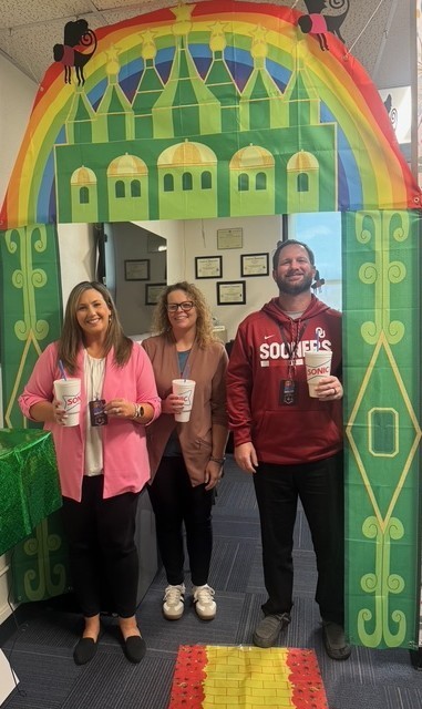 Three people in front of a backdrop of a castle with a rainbow arch. They are holding cups.