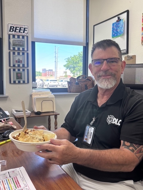 Man in glasses holds bowl of food. White sign on wall says "BEEF." Window in background.