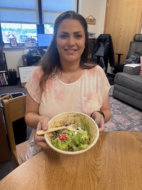 Woman seated at a table with a bowl of salad, smiling. Behind her, a desk and couch are visible.