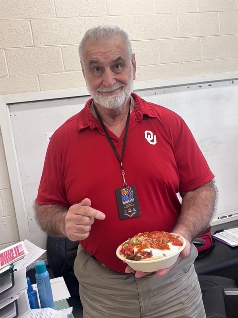 A man in a red shirt holds a bowl of food and points at it. A white board is behind him.