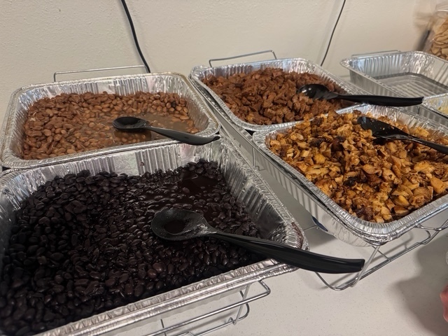 A table holds four aluminum pans filled with different foods: one with black beans, one with brown beans, and two with brownish-yellow food.