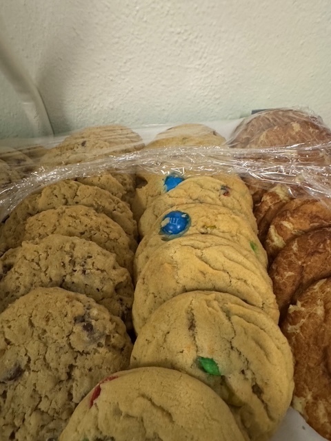 A collection of cookies, including chocolate chip, chocolate-coated, and M&M's cookies, arranged in a row and covered with plastic wrap.