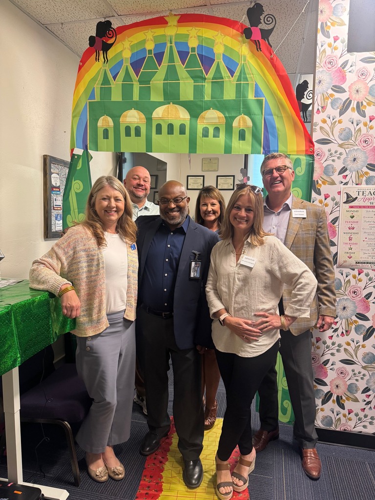 Five people stand in a room, three women and two men. They stand in front of a colorful arch with a rainbow and a castle.