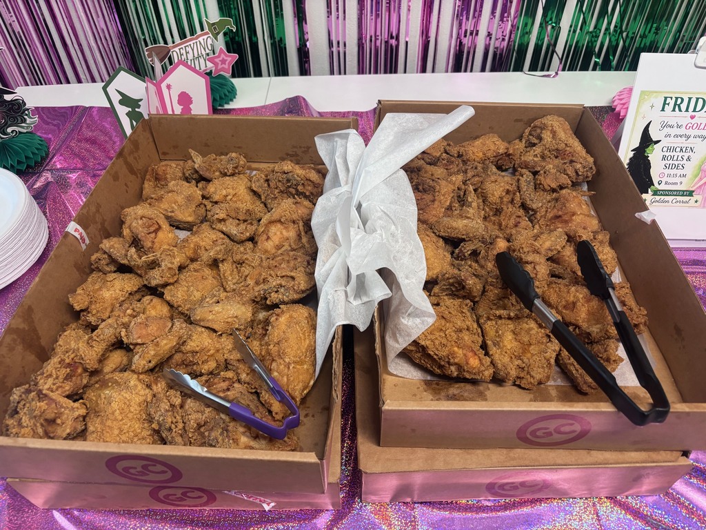 Two boxes of fried chicken on a purple table with a sign on the right. A stack of white plates on the left.