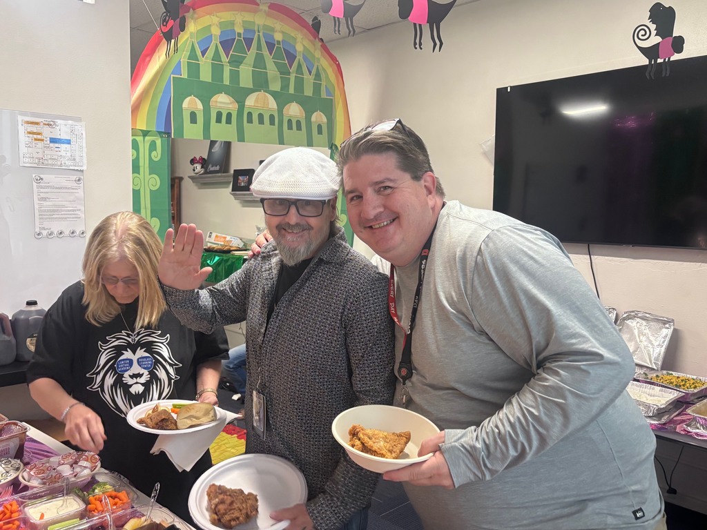 Three people at a buffet table with food. One person waves. A large painting is behind them.