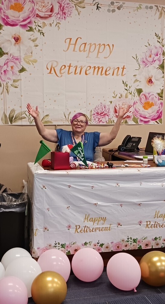 Person wearing glasses and a blue shirt celebrating retirement at a table with balloons and decorations.