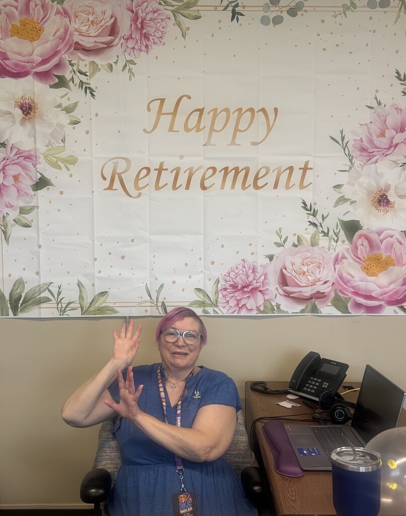 A woman in a blue dress sits at a desk, waving, with a retirement banner in the background.
