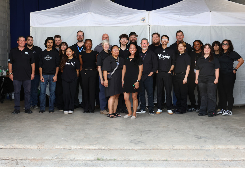 A group of people in black t-shirts and sneakers, some with lanyards, poses for a photo under white canopies.