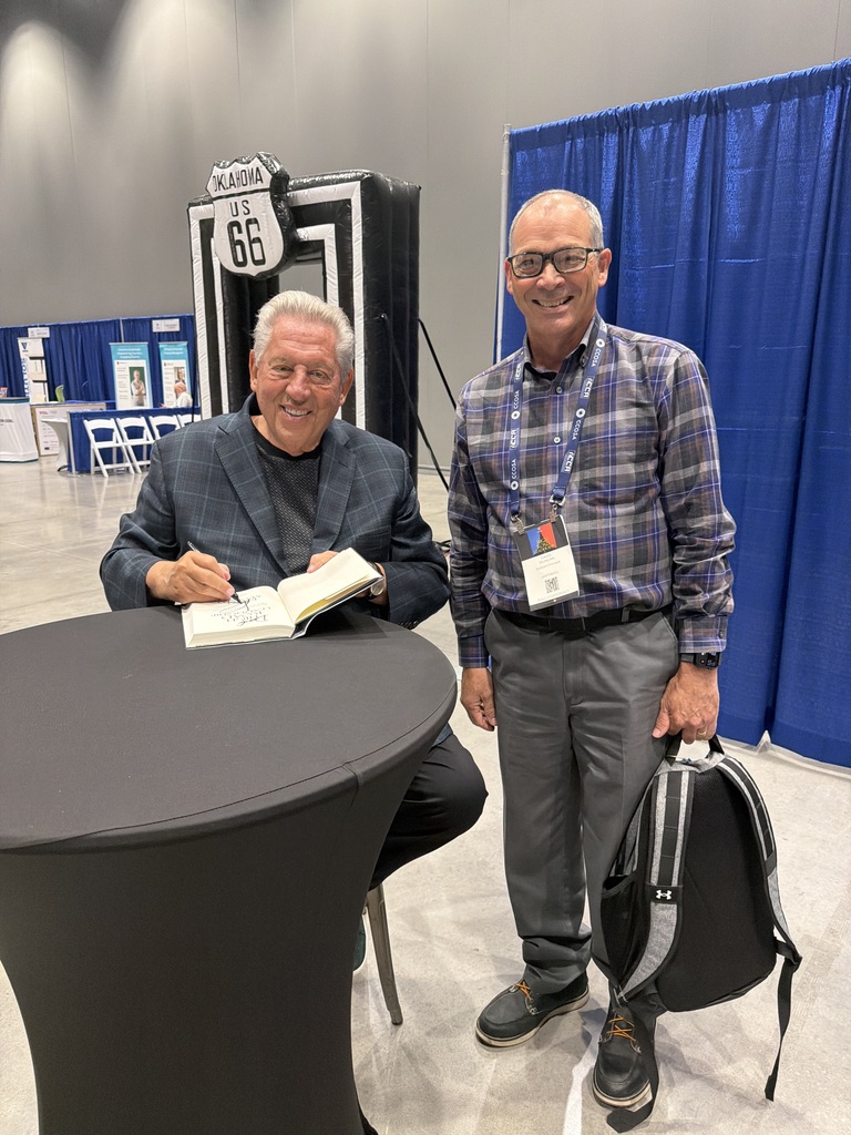 Two men posing. One signing a book and the other smiling.