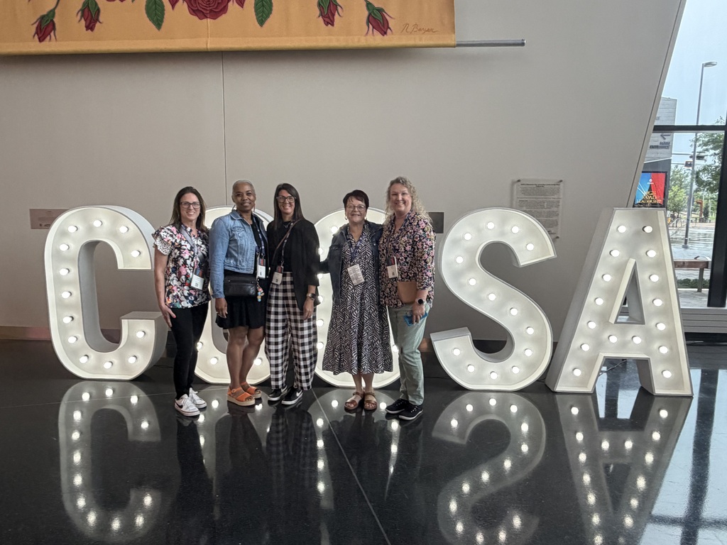 Five women in casual attire standing behind large letters spelling out "CONSA". A decorative wall backdrop behind them.
