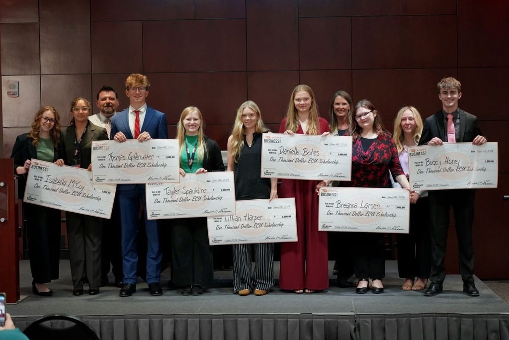 Group of people standing on a stage holding certificates with text, standing in front of a brown wall.