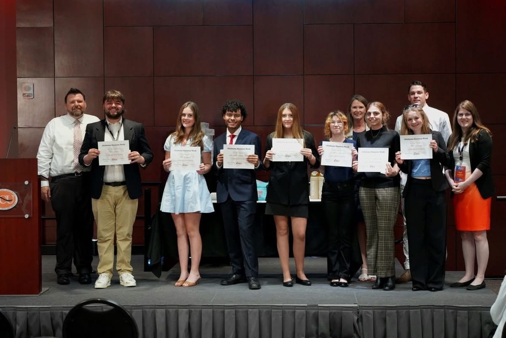 People stand on stage holding certificates. A podium is behind them. A dark backdrop is in the background.