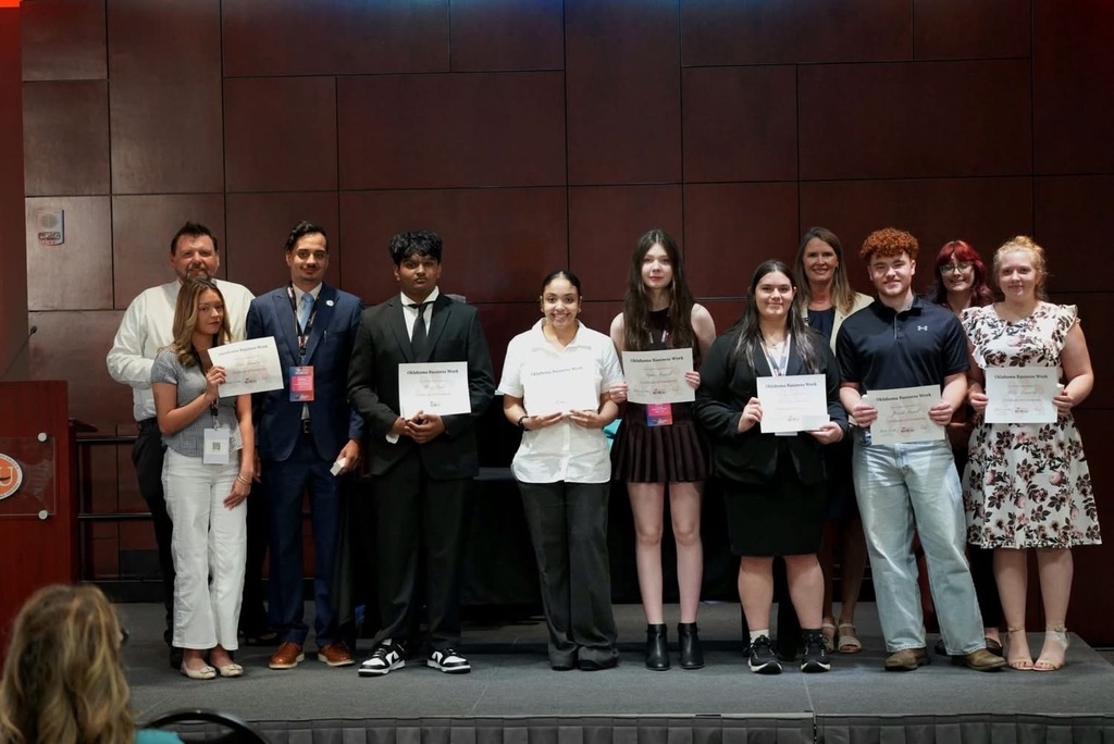 A group of people stand on a stage holding certificates, in front of a brown wall.
