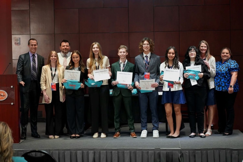 A group of students and adults stand on a stage holding certificates. They are dressed formally.