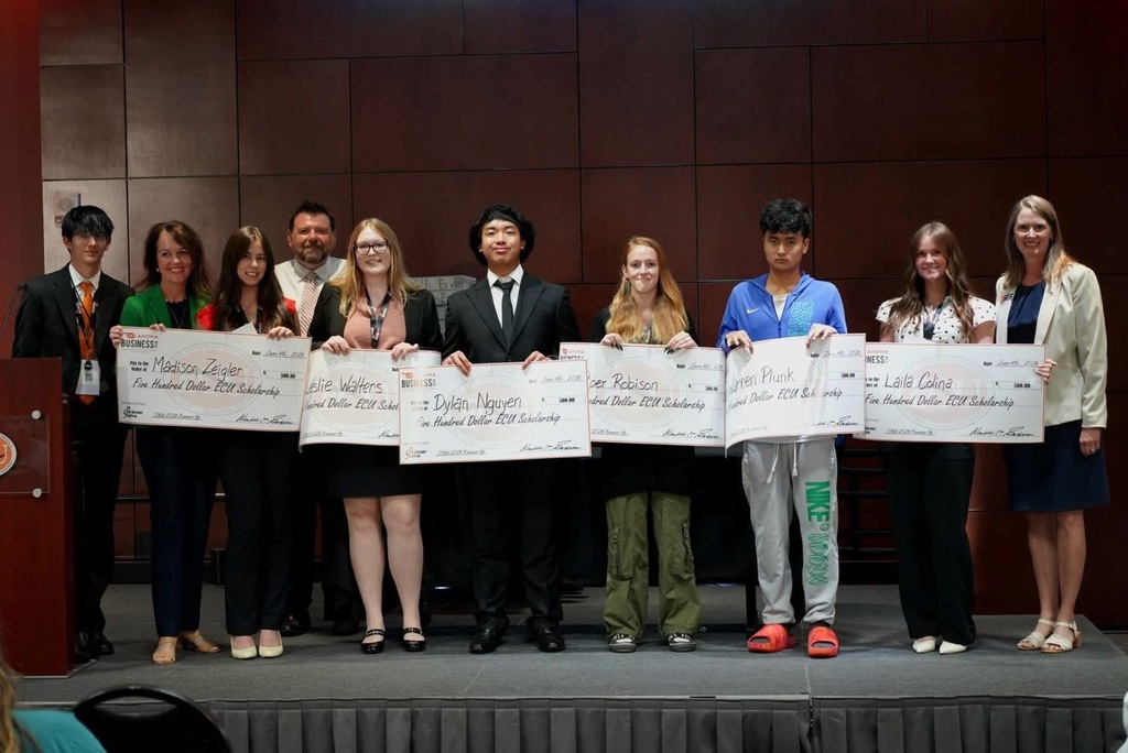 A group of people stand on a stage holding certificates and looking at the camera. They wear various outfits.
