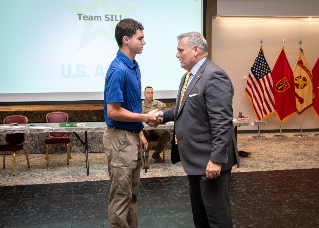 Two men shake hands in a room. A projector screen reads "Team SILL". Flags stand behind the men.