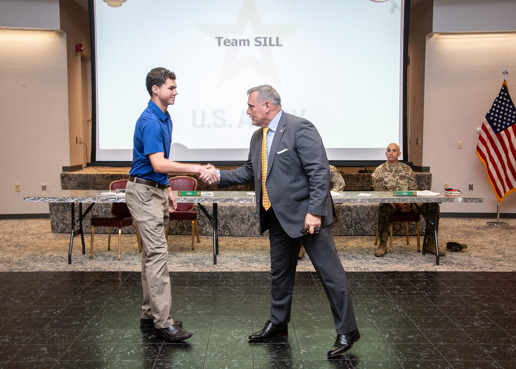 Two men shake hands on a stage. One wears a blue shirt, the other a gray suit. Behind them is a projector screen, a table, and a flag.