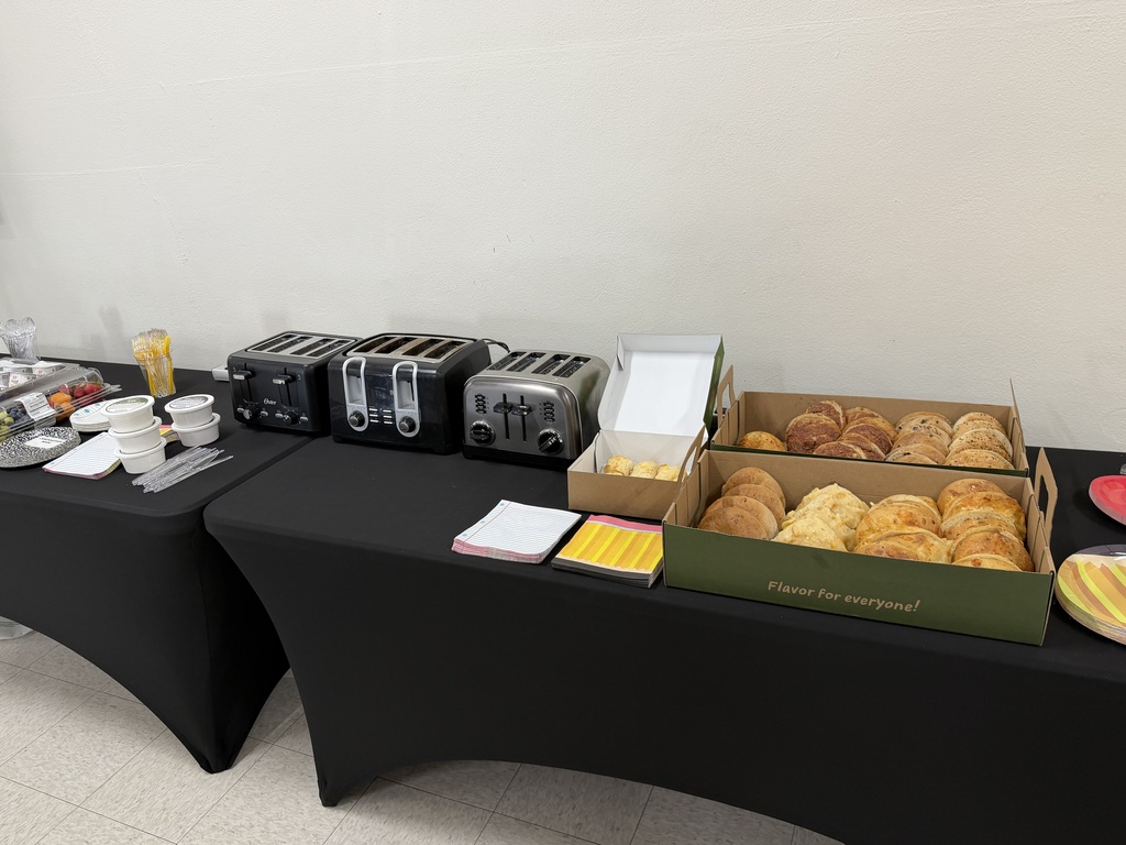 A table with black tablecloths holds bread in boxes, toasters, and other items. Behind the table is a plain white wall.