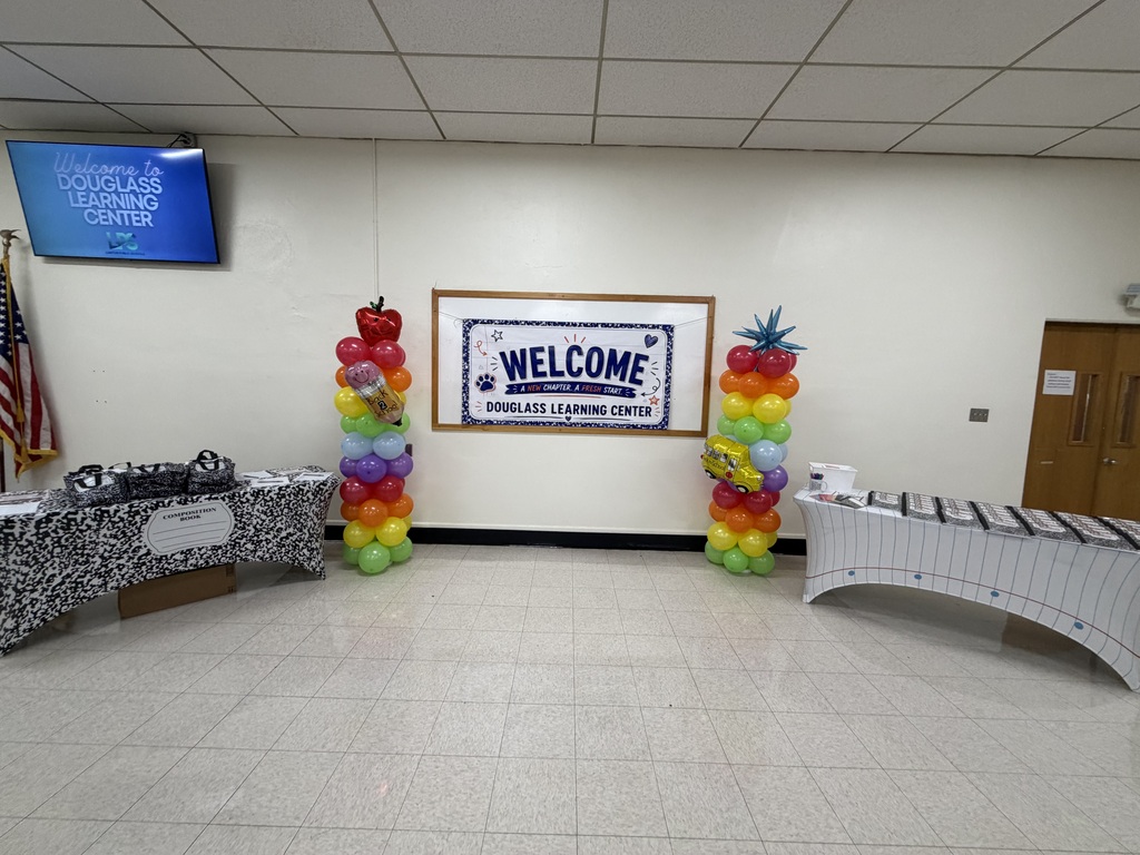 Empty room with white walls, two balloon stands, and a "Welcome" sign. Tables have black and white designs. Flag and TV on the wall.